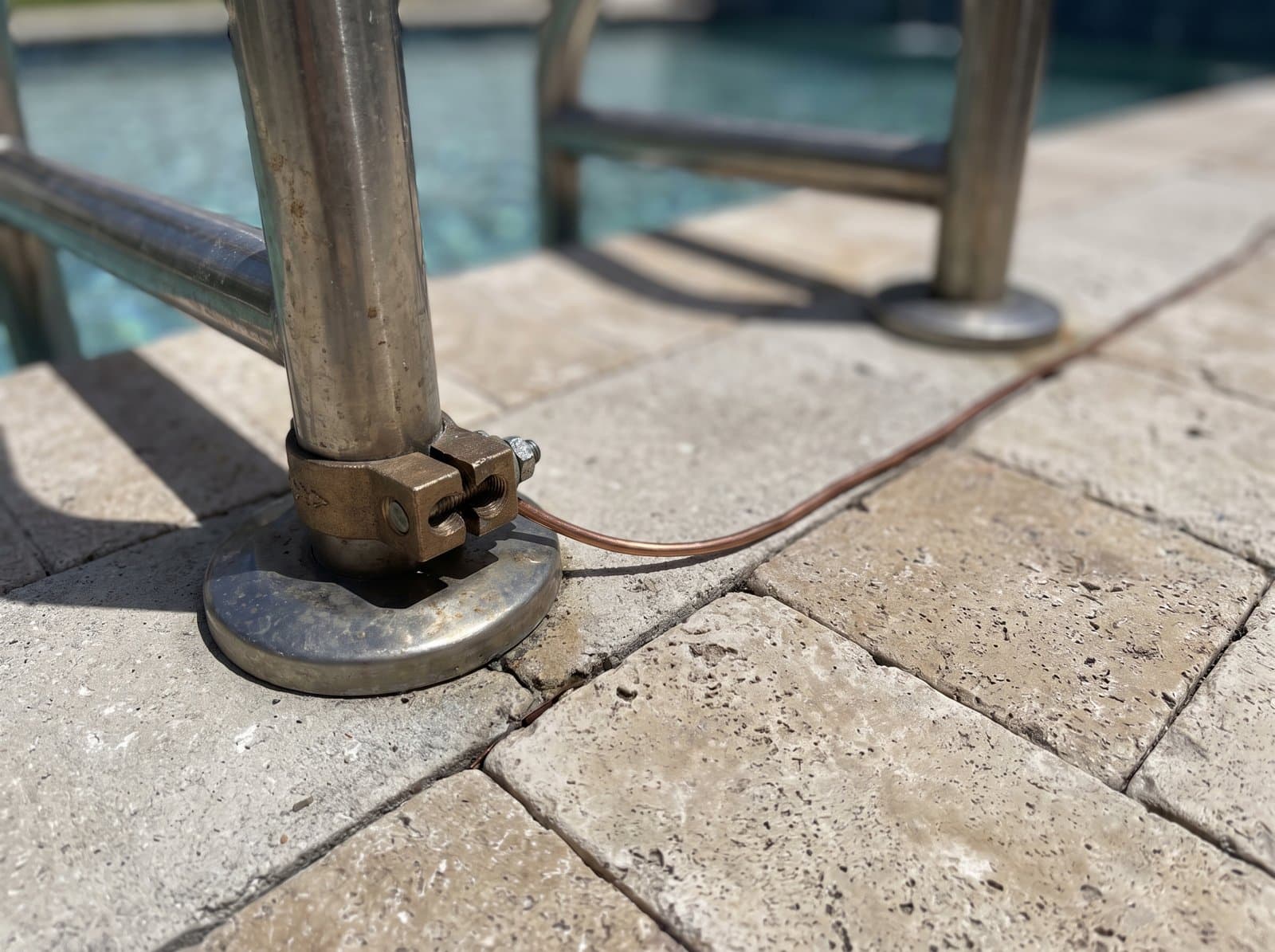 A bronze bonding lug clamped to a pool ladder rail with bare copper bonding wire across a travertine deck