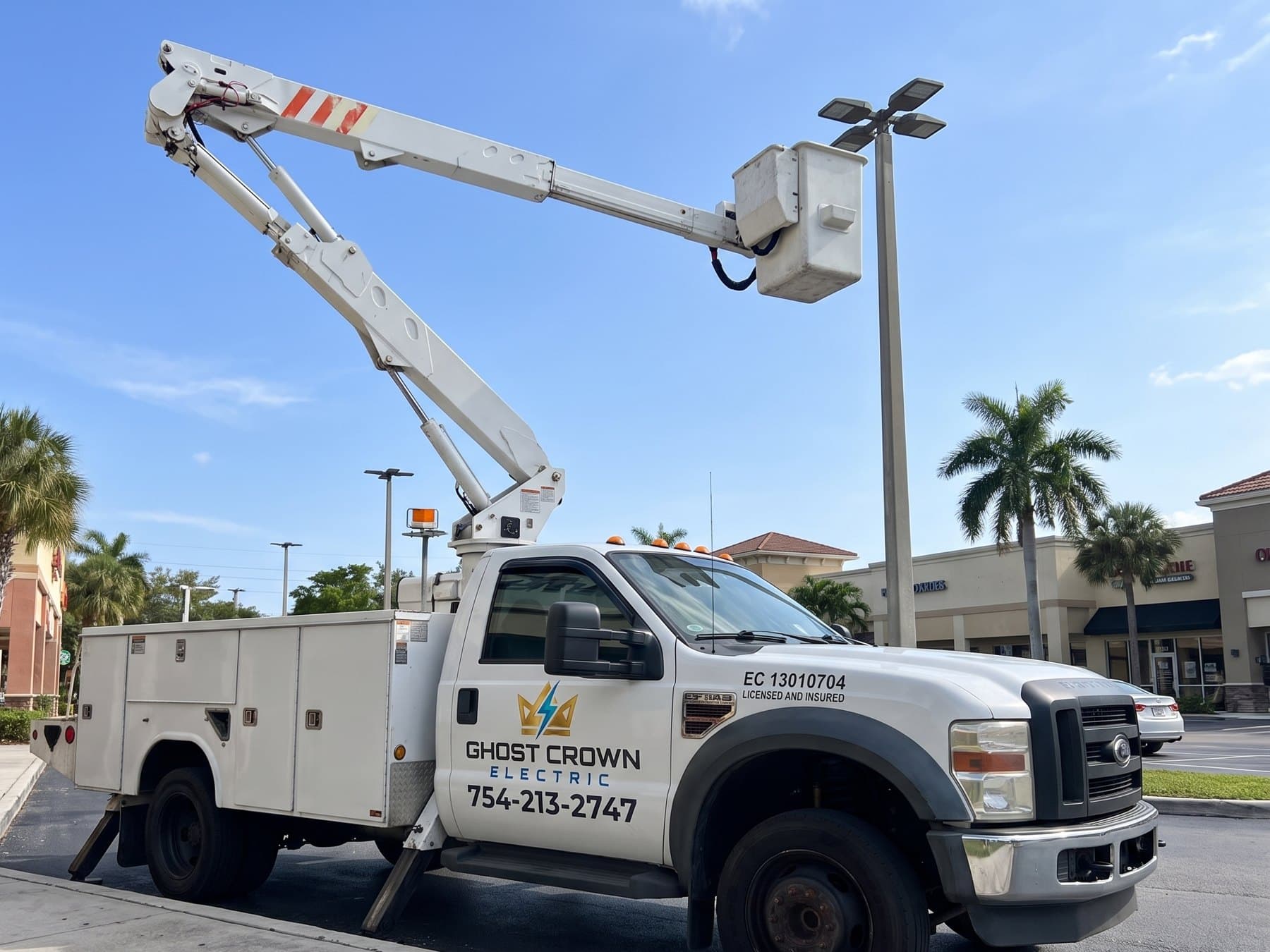 The Ghost Crown Electric bucket truck with its boom fully extended to a parking lot pole light at a South Florida plaza